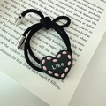Dettaglio dell'elastico per capelli nero con ciondolo a cuore Like posato su un libro - Stile Lettera Cuore / Nero (D-4810-4)