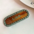Détail d'une barrette en acétate ambré avec bordure tressée verte et texte Like - Ambre et vert grisâtre