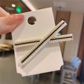 Dettaglio di un fermaglio a X dorato con perle e accenti neri su supporto di cartone - Croce Perla