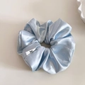 Detail view of light blue satin scrunchie showing glossy texture and a minimal woven label - Blue