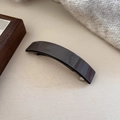 Dettaglio della molletta in acetato grigio scuro con finitura lucida su sfondo beige - Marrone