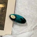 Detail of a dark green acetate oval hair clip with gold metal emblem on textured paper - Blue