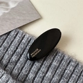 Glänzende schwarze ovale Haarspange mit Goldlogo auf grauem Strickgewebe - Schwarz