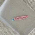 Close-up of a pink metal drop hair clip with a blue rectangular gemstone on a ceramic tray - Pink Waterdrop Clip (Blue Crystal)