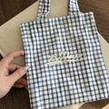 Detalle de bolso de lona a cuadros azules y blancos con texto Alline en mesa de madera - Bolso de Mano de Rayas Cruzadas Azules