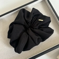 Close-up of black pleated fabric scrunchie with a small golden brand tag in a display tray - Black
