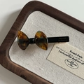 Detailed view of a tortoise shell bow hair clip with gold logo on dark wood tray - Tortoiseshell Blue Bow Duckbill Clip