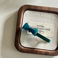 Detailed macro view of a teal acetate bow hair clip with gold logo on a wooden tray - Blue