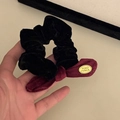 Detail eines schwarzen Samt-Scrunchies mit dunkelroter Cord-Schleife und goldenem Logo-Anhänger - Rote Schleife Goldener Anhänger Schwarzer Haargummi