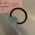 Detail shot of a black elastic hair tie featuring a light green resin charm with pink smiley face - Green Bear Hair Tie