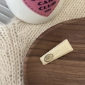 Primer plano de pinza de pelo de acrílico crema con logo dorado en bandeja de madera - Blanco Roto