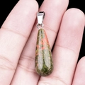 Colgante de gota en unakita natural con patrones verde y rosa sostenido por dedos - Unakita