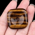 Polished tiger eye square pendant with central hole held by hand on black background - Tiger's Eye