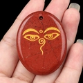 Oval red jasper pendant with golden Buddha eye pattern held in hand against black backdrop - Red Jasper