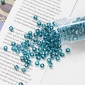 Close-up of 4mm teal glass seed beads with silver inner core pouring from a tube - Peacock Blue