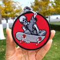 Parche bordado redondo rojo con calavera skater sostenido sobre un fondo desenfocado - Estilo Esqueleto con Monopatín (PE5778CT)