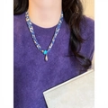 Primo piano di una collana in tessuto floreale blu con pendente a goccia su maglia viola. - Floreale / Blu