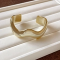 Dettaglio ravvicinato di un bracciale rigido dorato a onde geometriche su vassoio. - Stile 35 - Onda Dorata