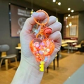 Close-up of heart-shaped orange quicksand keychain with glitters, pearls and small bell - Orange