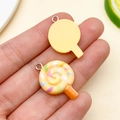 Close-up of resin lollipop charms held in hand showing colorful spiral details and metal ring - Yellow Resin Lollipop (24573) / 26x17x5mm