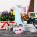 Detail of Mother's Day gnome doll holding a pink Love Mom heart in a cozy home setting - Blue