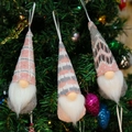 Dettaglio di tre gnomi natalizi in maglia appesi a un albero con luci calde e festive - Stile Rosa Grigio (C)
