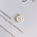 Dettaglio del ciondolo rotondo dorato con lettera S in smalto bianco su carta - Stile S