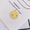 Dettaglio di un ciondolo emoji giallo con viso che bacia su un biglietto EMBRACE - 10# Bacio a Sinistra