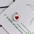 Macro shot of white enamel heart charm with gold rim on a minimalist display card - Red