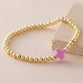 Golden beaded bracelet featuring a pink cross charm displayed on a textured stone base - Purple
