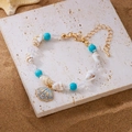 Perlenarmband mit blauem Emaille-Muschelanhänger und Goldakzenten auf hellem Sandbett - Blau Strass Muschel