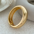 Close-up of polished gold-tone geometric bangle with spring hinge mechanism on a table - Chunky Tubular Bangle (2609) / Gold