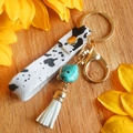 Llavero boho con estampado de vaca, piedra turquesa y borla blanca en fondo de madera - Estampado de Vaca (Galvanoplastia de Oro 1)