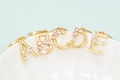 Makro-Detail von vergoldeten A-E Buchstaben-Anhängern mit Zirkonia-Besatz auf weißer Keramik - Buchstabe A / Gold