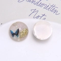 Detalle de amuleto de resina con mariposa azul, rosa dorada y purpurina con reverso plano blanco - Estilo Redondo