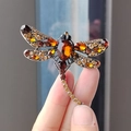 Broche libellule vintage avec strass ambrés, tenue à la main sur un fond bleu doux et flou - Petit Brun (XZ0207-6)