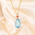 Close-up of golden necklace with red bead and light blue teardrop pendant on satin - Necklace