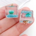 Hand holding two luminous square resin pendants with blue and green dried flowers inside - Blue Green Cube (19178)