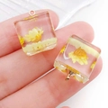 Handheld view of a square resin pendant with yellow dried flowers and gold ring hook - Yellow Cube (19181)