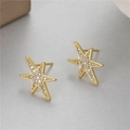 Detalle de pendientes de estrella dorados con circonitas sobre una superficie mate gris - Estilo de Estrella Hexagonal Asimétrica (H-10203)