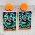 Detail of tarot card style earrings featuring a skull hand holding coffee beans on gray surface - The Coffee (JE1040A) / Orange