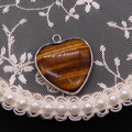 Detailopname van hartvormige tijgeroog hanger met zilveren rand op kant en parel achtergrond - 26x28mm, 7g / Tijgeroog