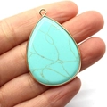 Waterdrop-shaped turquoise stone pendant with golden metal border held in hand - Blue Turquoise