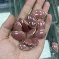 Hand met gepolijste druppelvormige roze natuursteen hangers met gouden metalen randen - Rode Houtnerfsteen