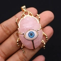 Detailaufnahme des handgehaltenen Gold-Skarabäus-Anhängers mit rosa Stein und blauem Auge - Rosenquarz
