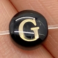 Macro shot of a black round sea shell bead with a golden letter G for DIY jewelry - Letter G / 8mm