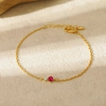 Detail of gold chain bracelet featuring red gemstone and clear zircon with lobster clasp - July