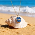 Goldene Evil Eye Halskette mit blauem Emaille-Anhänger auf einer Muschel am Strand - Schutzauge - Dunkelblau