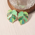Golden Monstera leaf earrings with green and blue enamel on a light wood background - Green