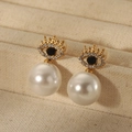 Macro shot of gold crown eye earrings with large pearls on beige textured jewelry box - Evil Eye Style